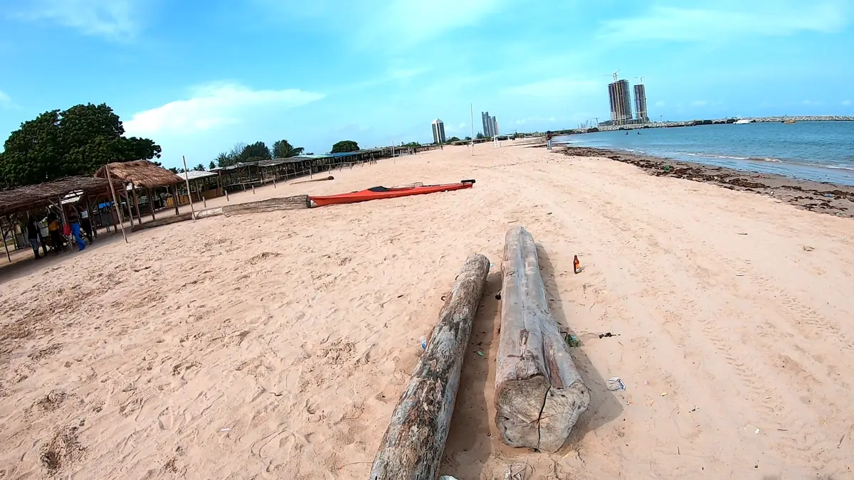 Sandy beach front at Tarkwa Bay, Lagos, with driftwood logs, a red kayak, thatched shelters and high-rise towers across the water