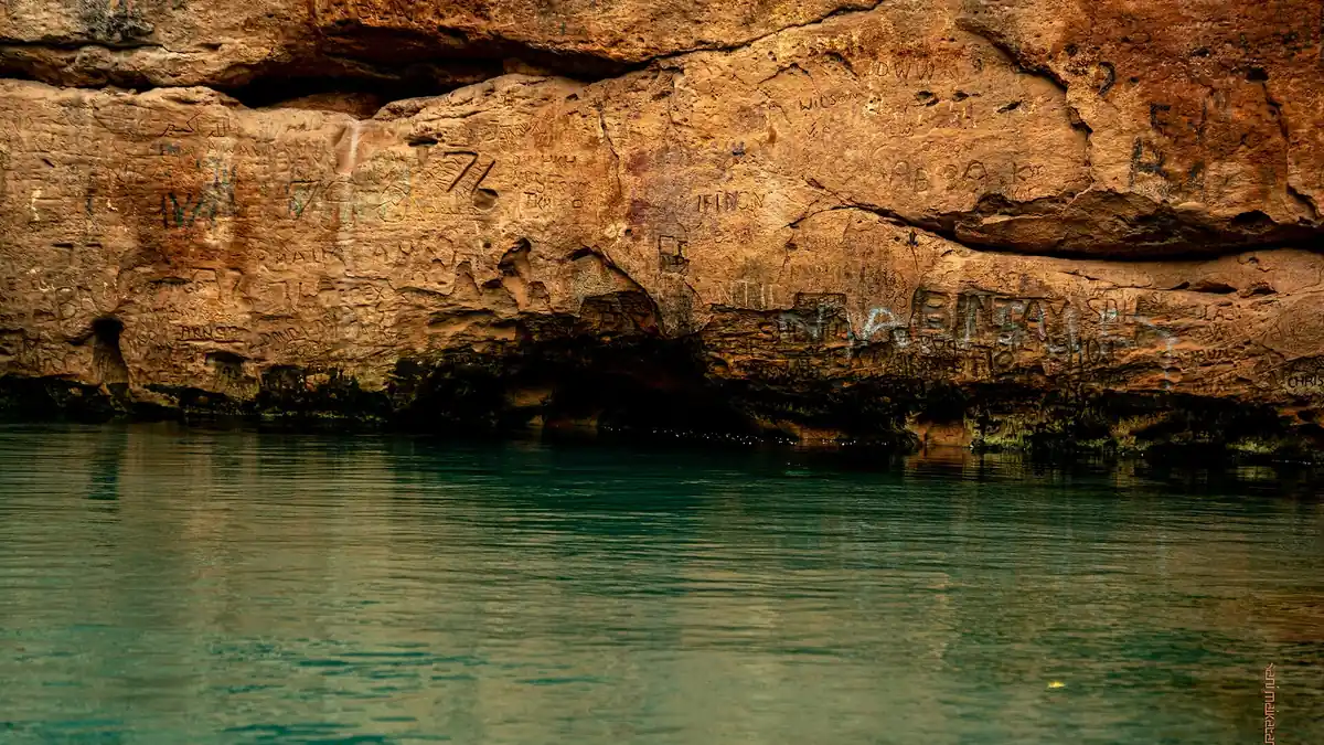 Clear green water of Wikki Warm Spring in Yankari Game Reserve beneath a sandstone cliff carved with graffiti