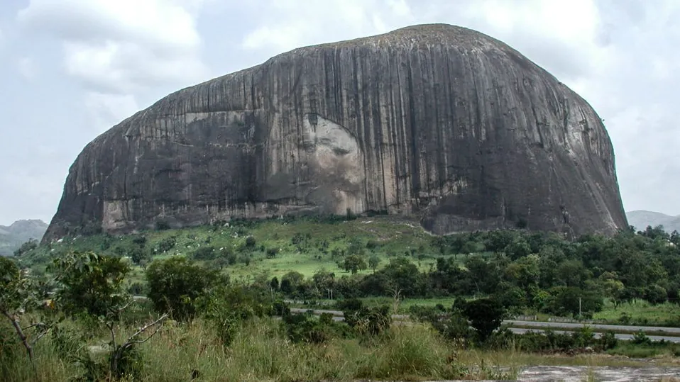 Zuma Rock, a huge dark granite monolith, rising above green bush and a road under a cloudy sky